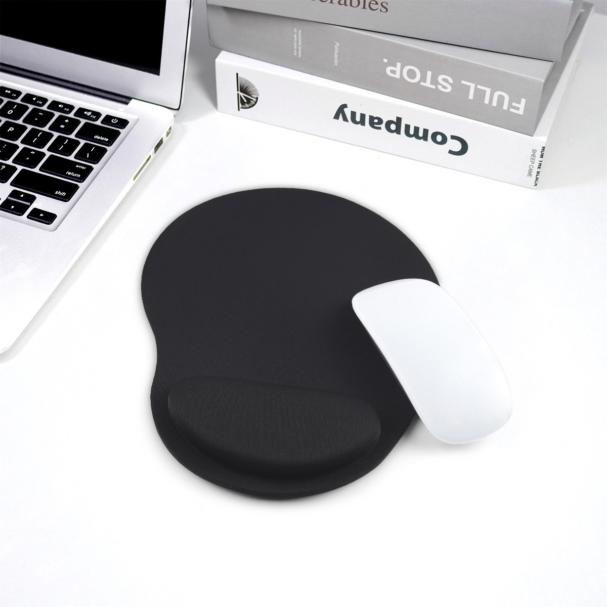 Eine weiße Maus liegt auf einem schwarzen ergonomischen Mauspad neben einem Laptop und Büchern.