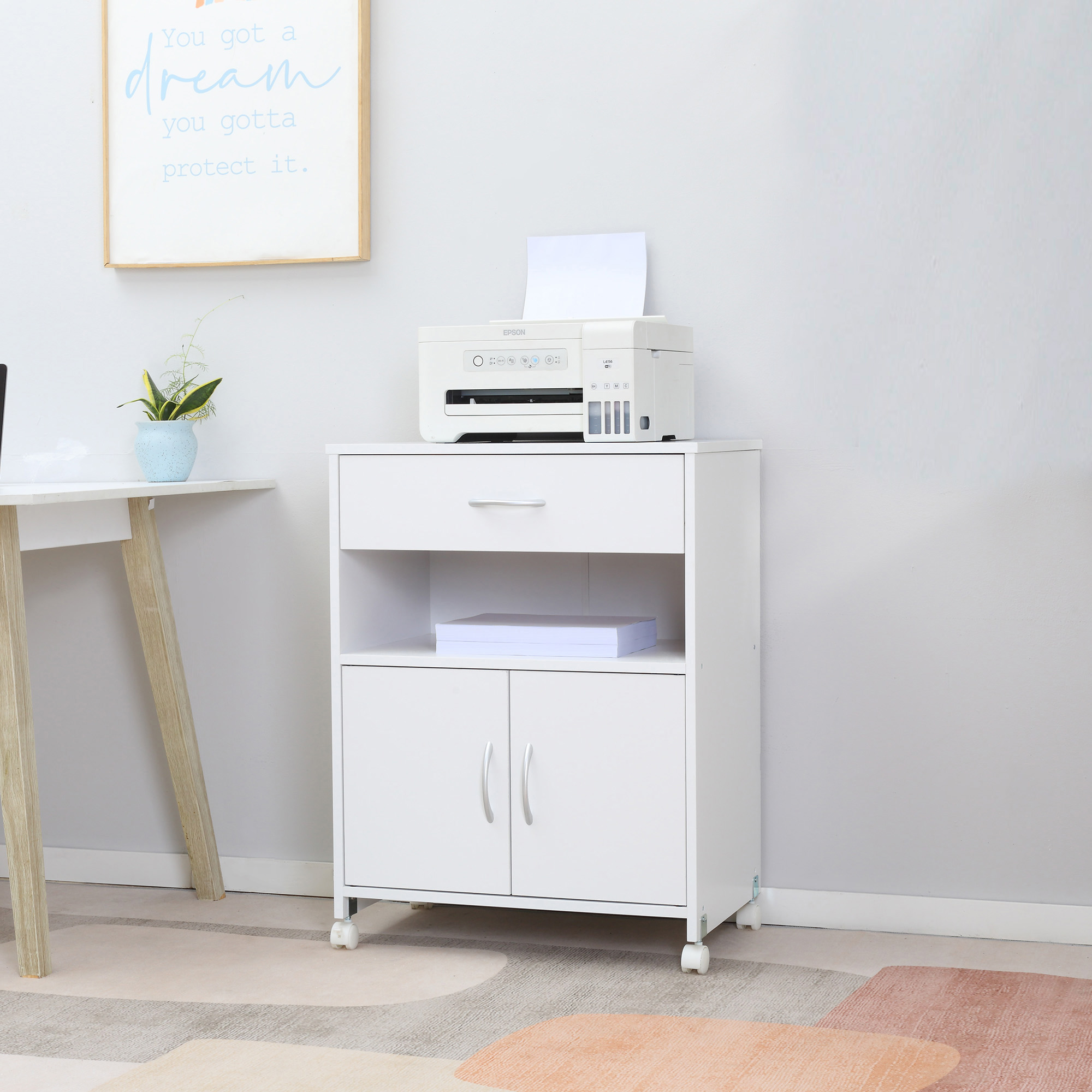 Stampante bianca su mobile bianco con cassetto e ante. Sopra è appeso un quadro incorniciato.