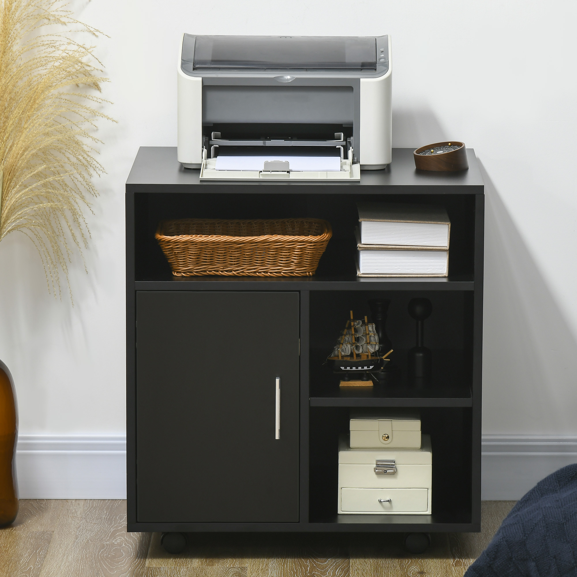 Carrello nero su ruote con stampante, cesto, libri e oggetti decorativi.