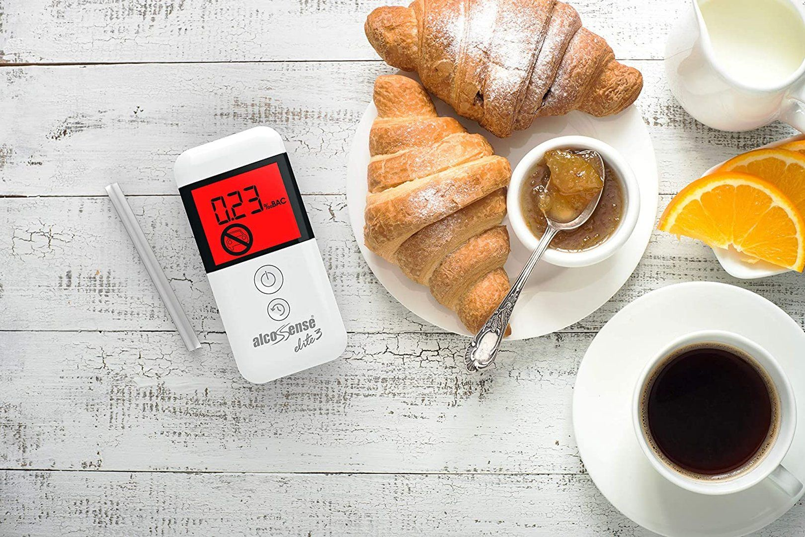Alkomat pokazujący 0.23% BAC leży obok croissantów, dżemu, kawy i pomarańczy.