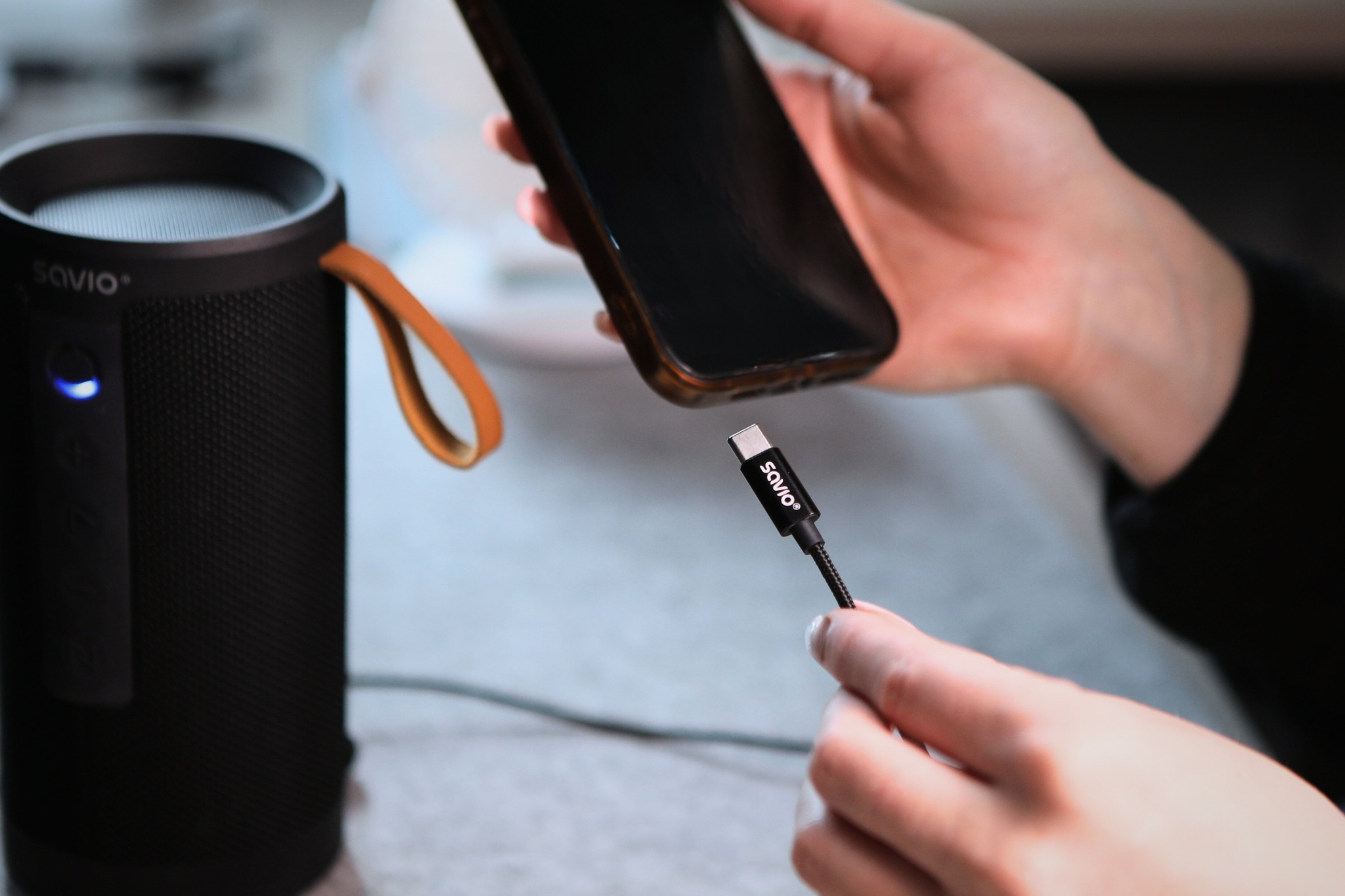 A hand holds a charging cable near a black speaker and a smartphone.