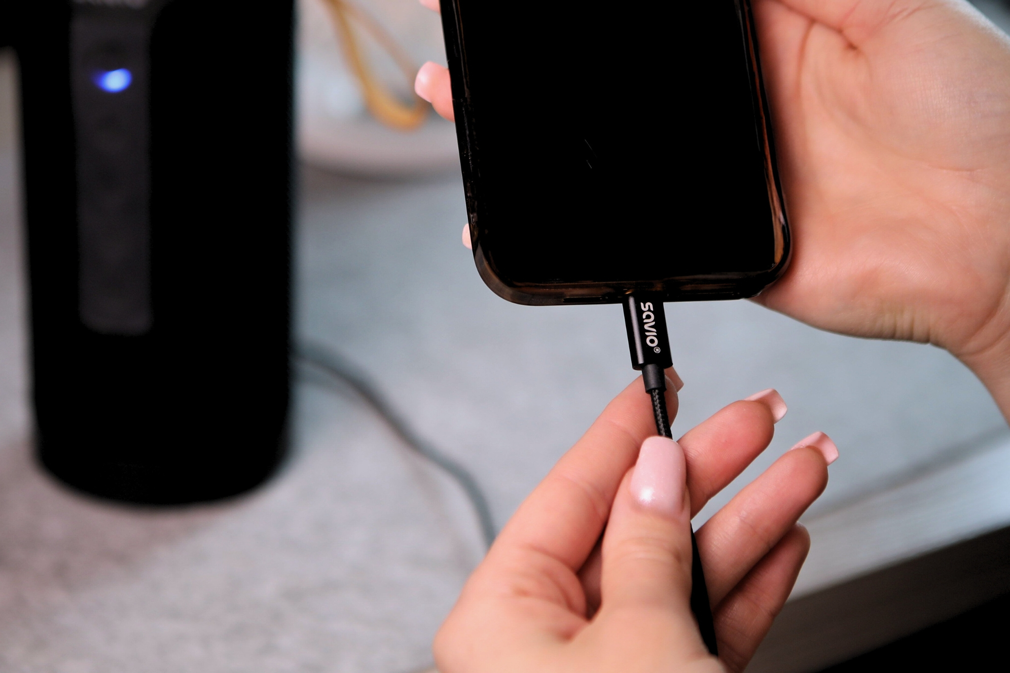 A hand plugs a Savio cable into a black smartphone. A black speaker is in the background.