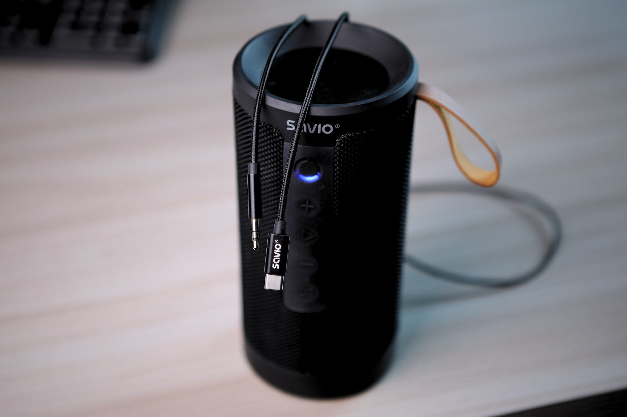 Black Savio Bluetooth speaker with cable and USB-C connector, on a wooden table.