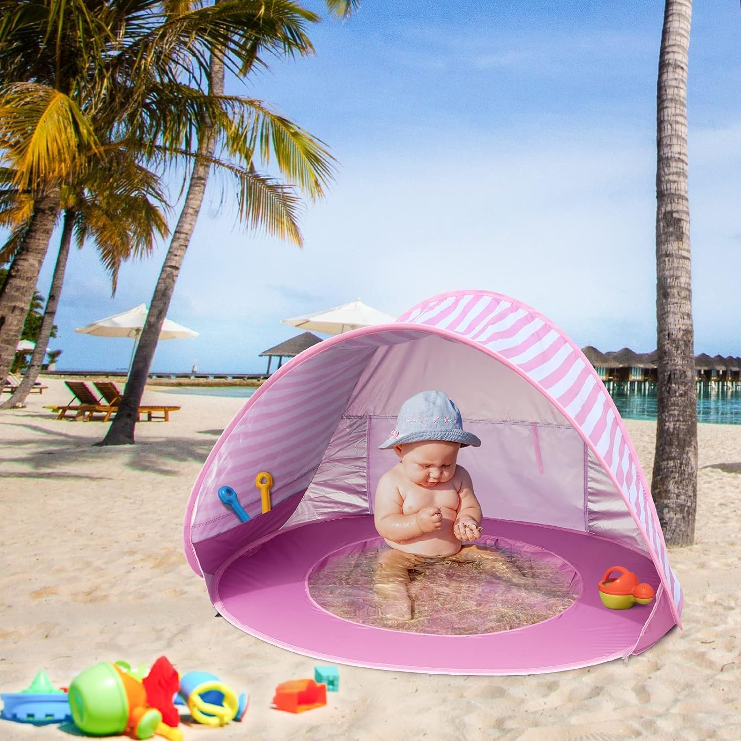 Baby sitzt in pinkem Zelt am Strand mit Spielzeug. Palmen und Ozean im Hintergrund.