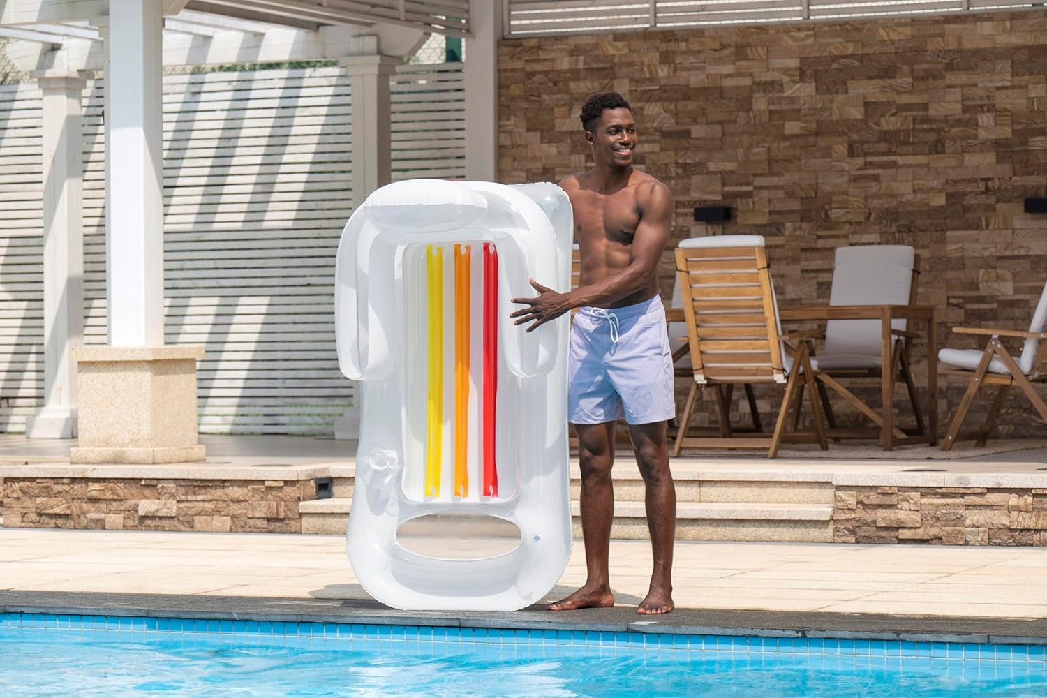 Ein Mann steht am Pool und hält eine weiße Luftmatratze mit bunten Streifen.