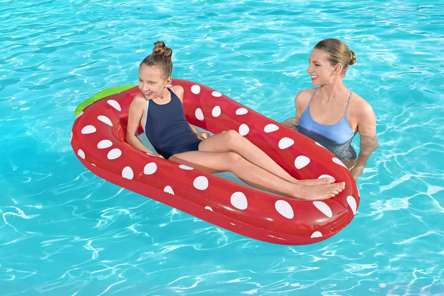 Ein Mädchen sitzt auf einer roten Erdbeer-Luftmatratze im Pool, eine Frau steht daneben.