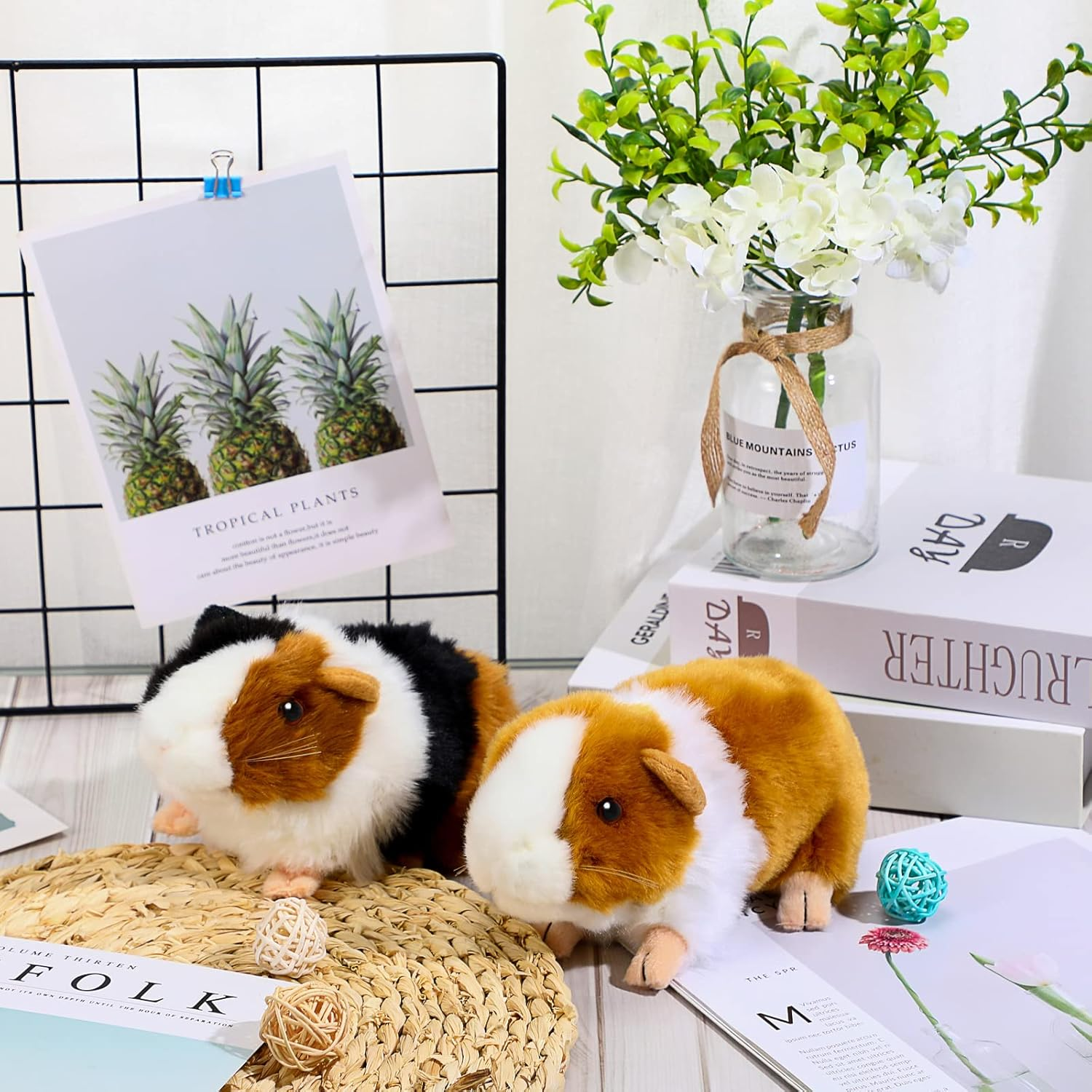 Zwei Plüschmeerschweinchen mit drei Ananas auf einem Bild, Blumen und Bücher im Hintergrund.