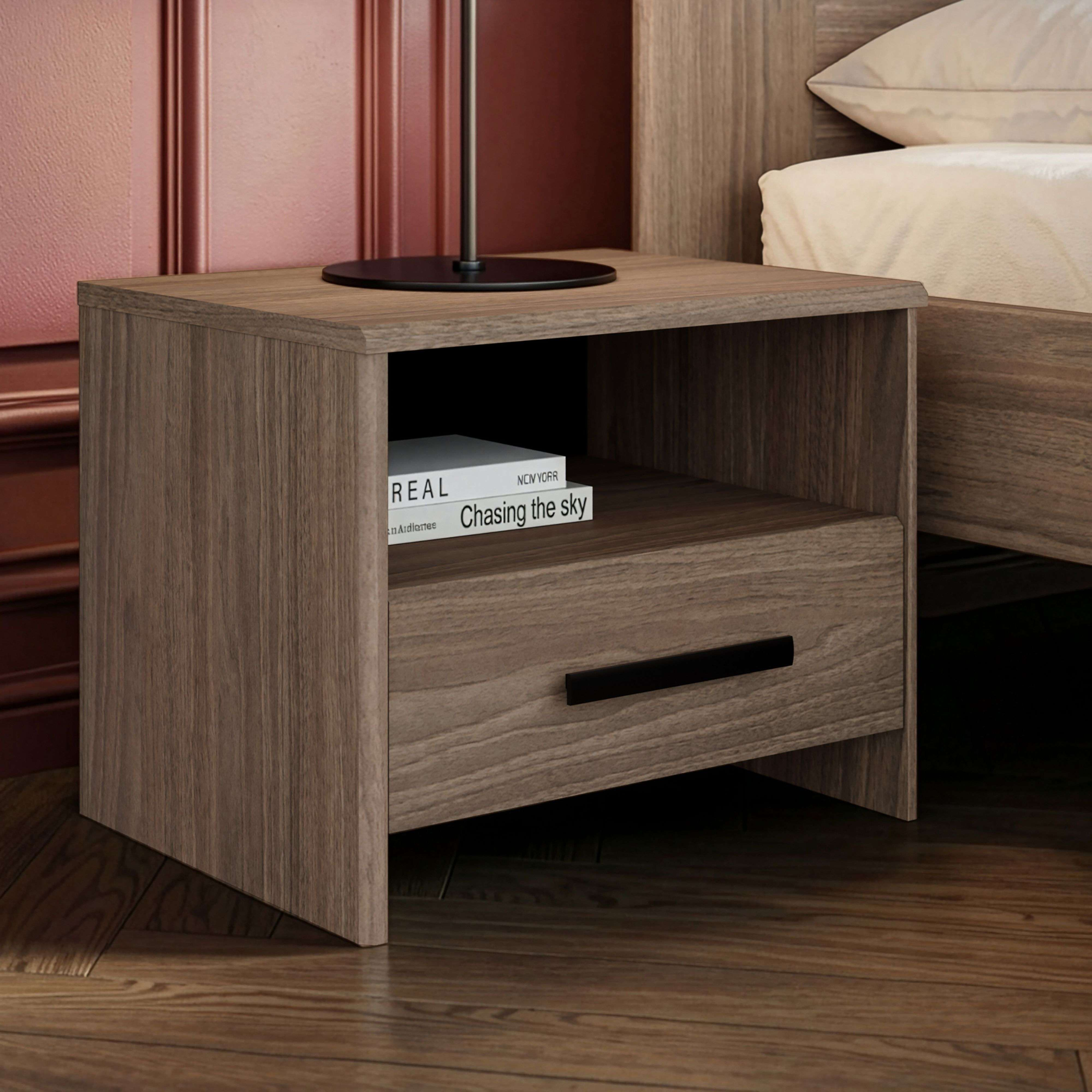 Una mesita de noche de imitación madera con cajón y estante abierto, junto a una cama.