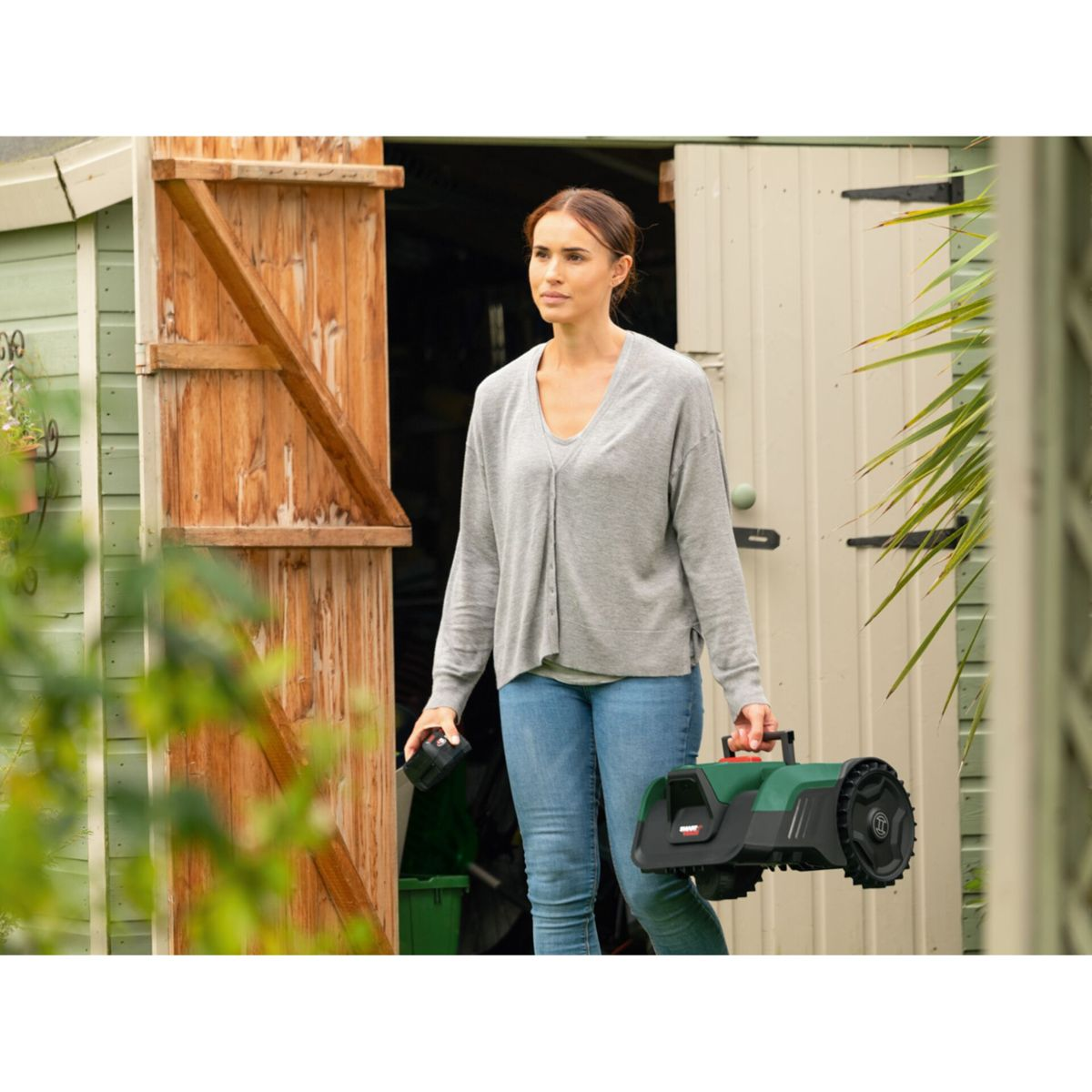 Eine Frau in Jeans und grauem Pullover trägt einen Mähroboter und Akku.