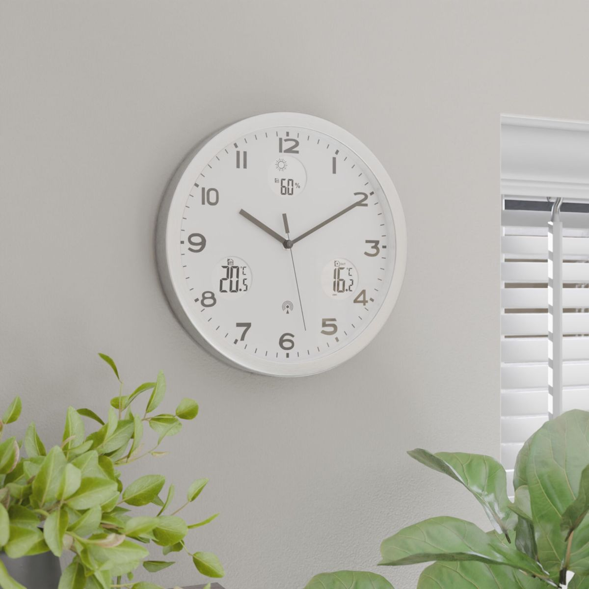 Eine weiße Digitaluhr an einer grauen Wand zeigt Uhrzeit, 60 % Luftfeuchtigkeit, 20,5 °C Innentemperatur, 16,2 °C Außentemperatur.