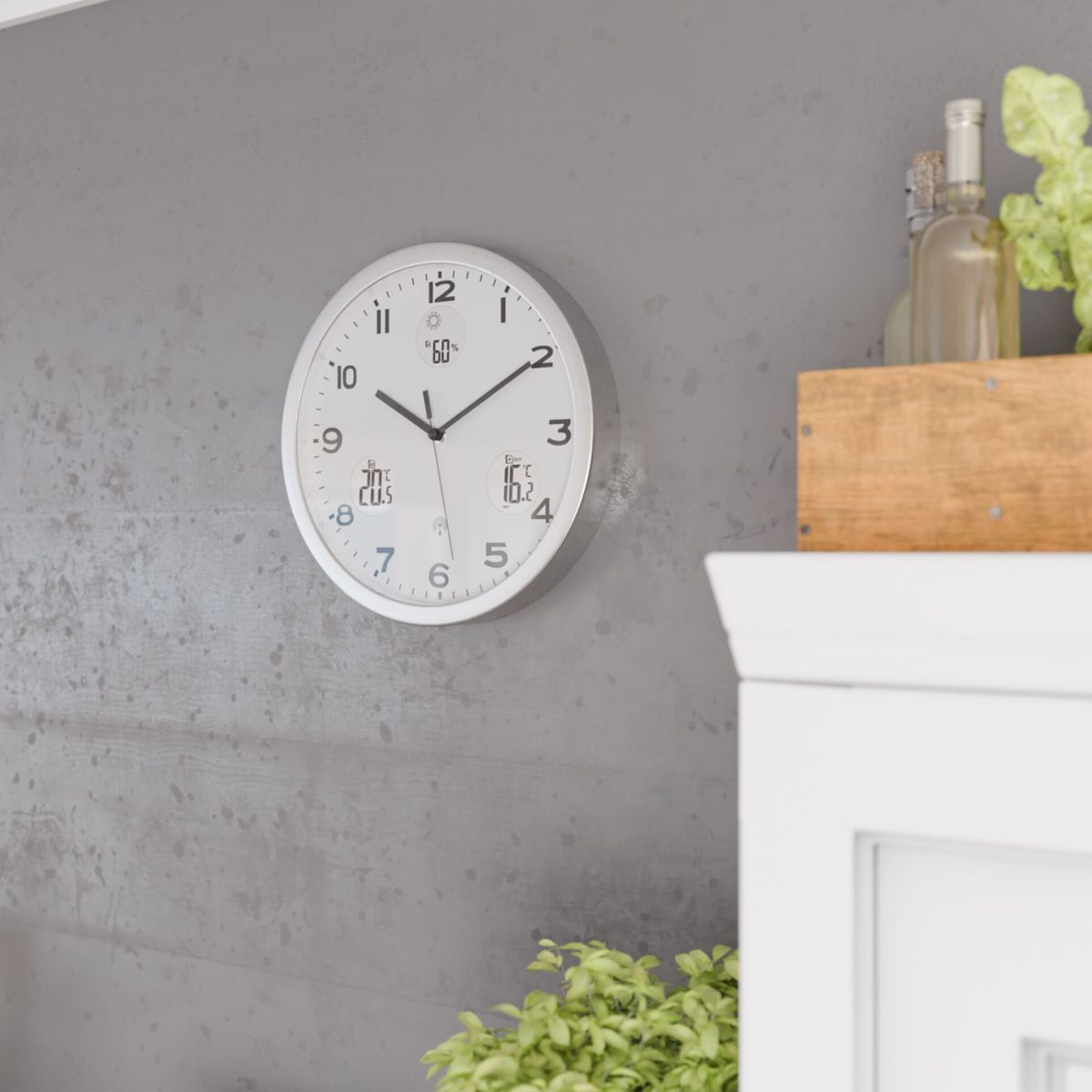 Eine weiße Wanduhr mit Temperatur- und Feuchtigkeitsanzeige hängt an einer Betonwand.