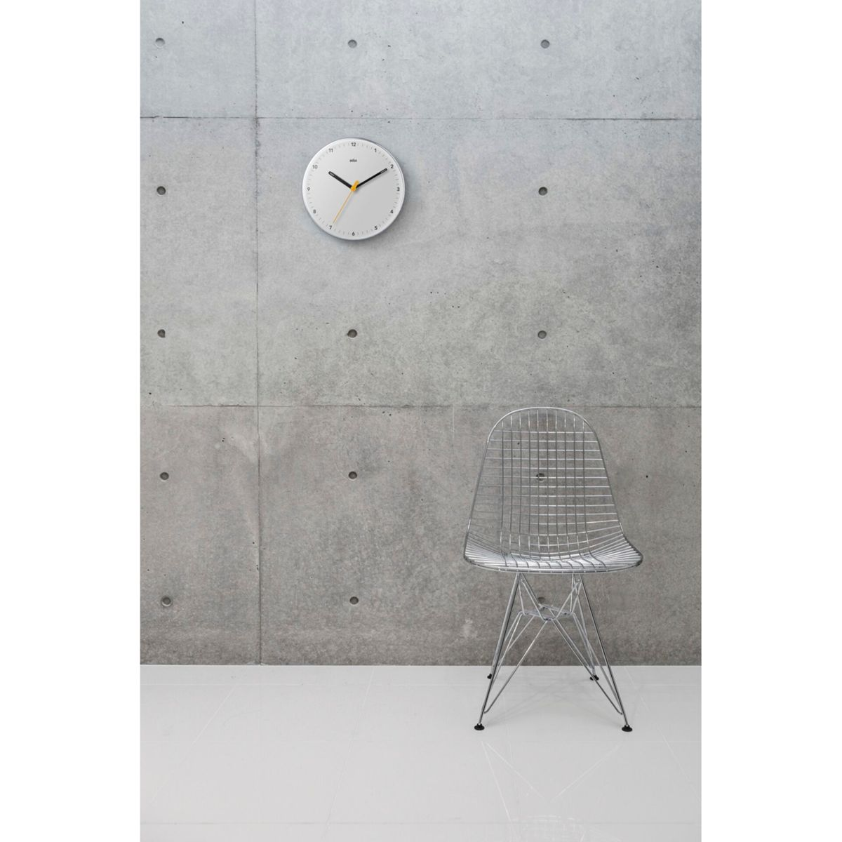 Eine weiße Wanduhr mit gelben Zeigern hängt an einer Betonwand über einem silbernen Drahtstuhl.