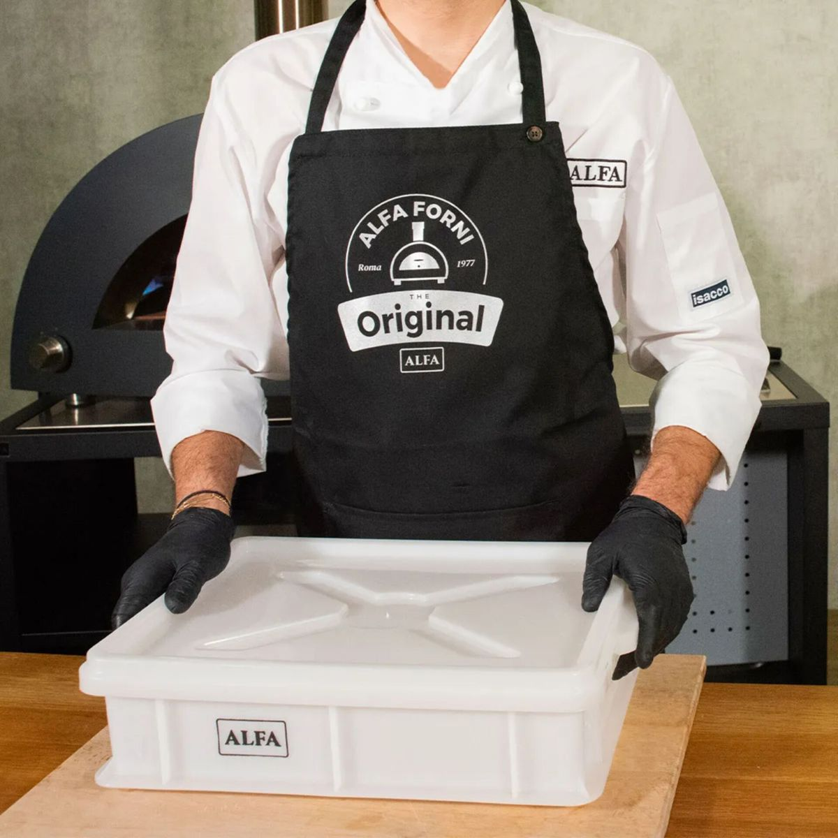 Koch in schwarzer Schürze mit "ALFA FORNI"-Logo hält weiße Teigbox vor Pizzaofen.