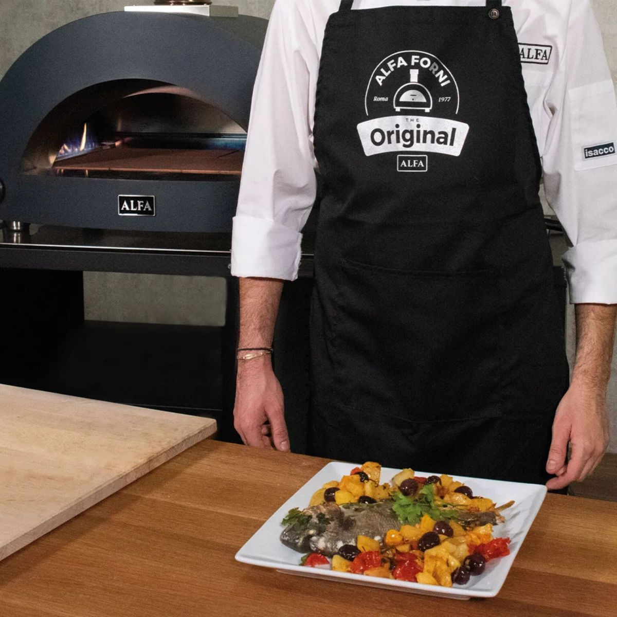Ein Koch in schwarzer Schürze präsentiert gebackenen Fisch mit Gemüse und Oliven.
