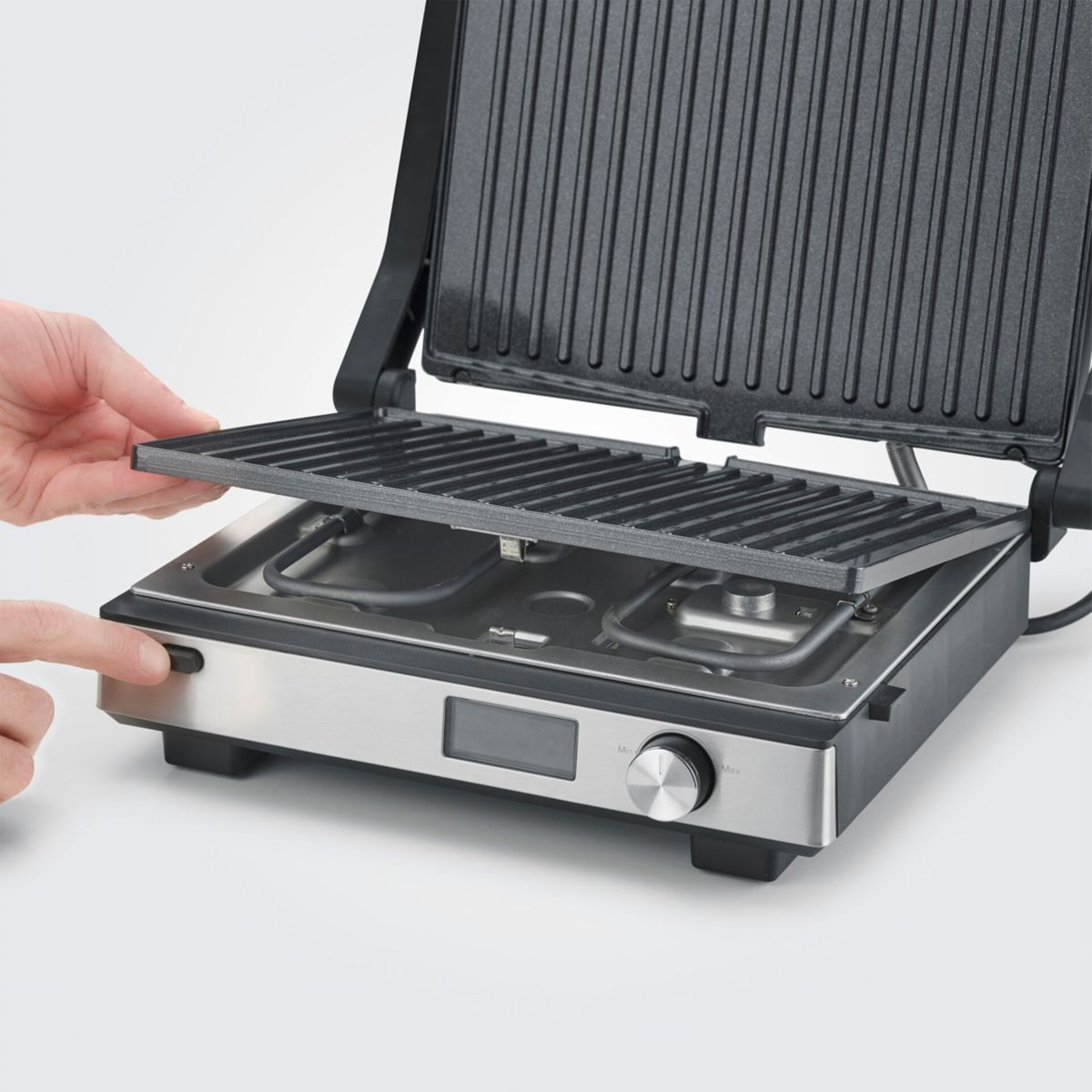 Ein Mann wechselt die Grillplatte eines Kontaktgrills aus. Metallgehäuse, schwarzer Griff.