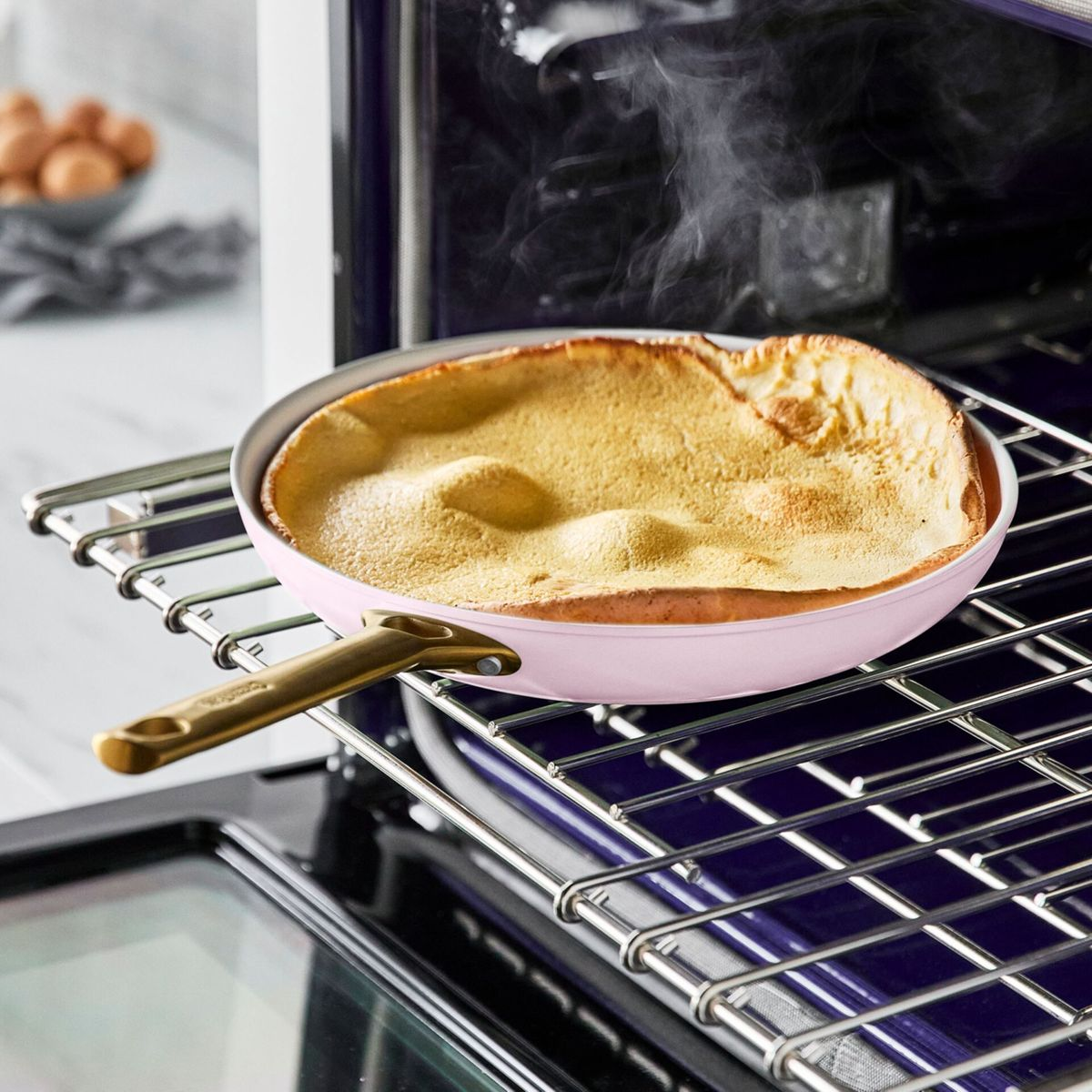 Ein hellrosa Pfannkuchen backt in einer Pfanne auf einem Ofenrost. Rauch steigt auf.