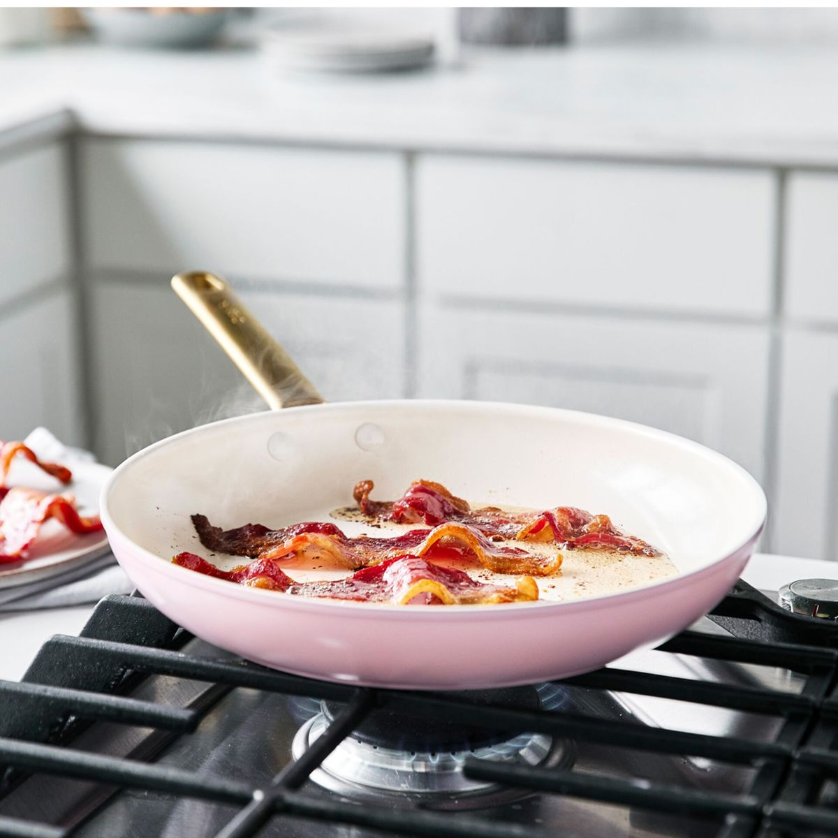 Bacon brät in einer rosa Pfanne auf einem Gasherd. Dampf steigt auf.