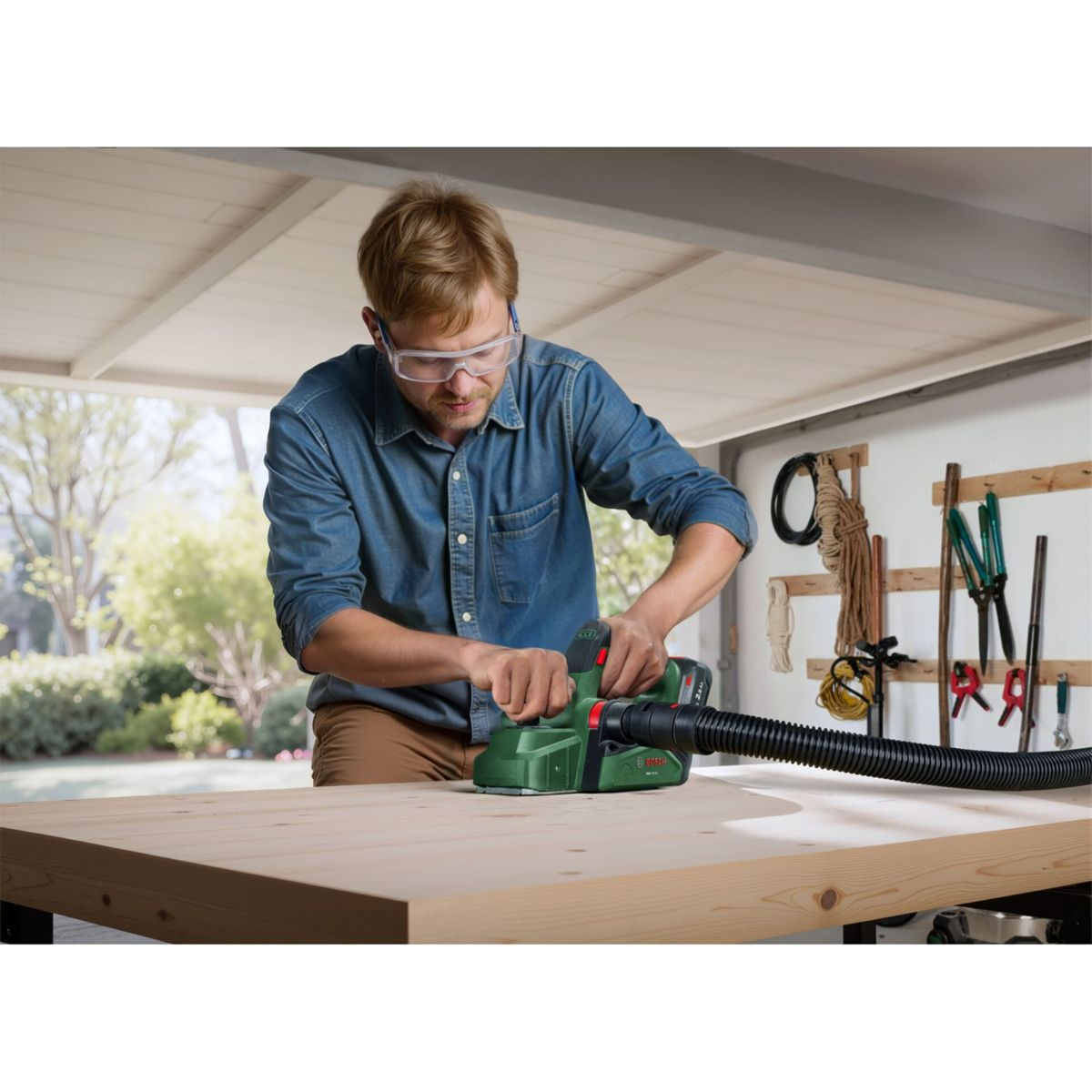 Mann in blauem Hemd und Brille benutzt grünen Schleifer auf Holz. Werkzeug hängt an Wand.