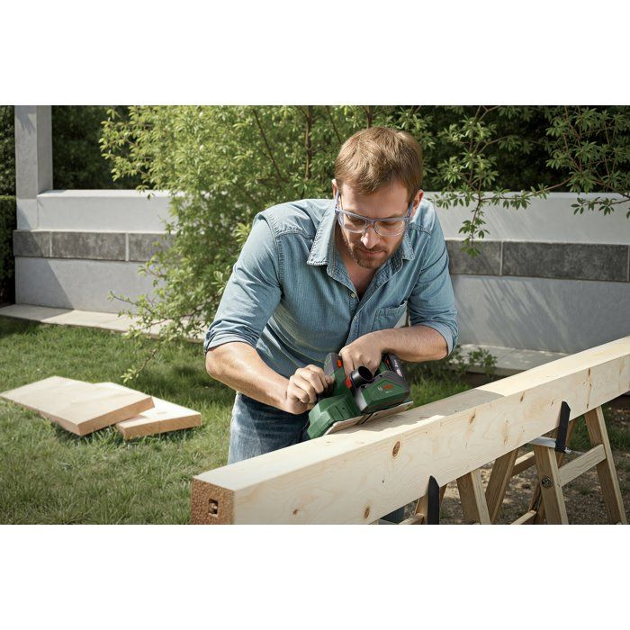 Ein Mann hobelt mit einem grünen Elektrowerkzeug einen Holzbalken im Garten.