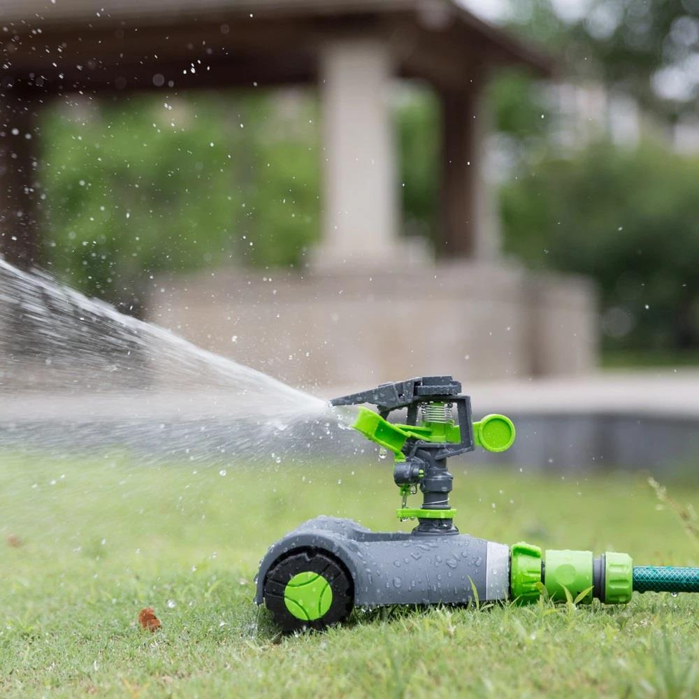 Tekerlekli çim sprinkleri yeşil çimlere su püskürtüyor. Arkada bir köşk var.