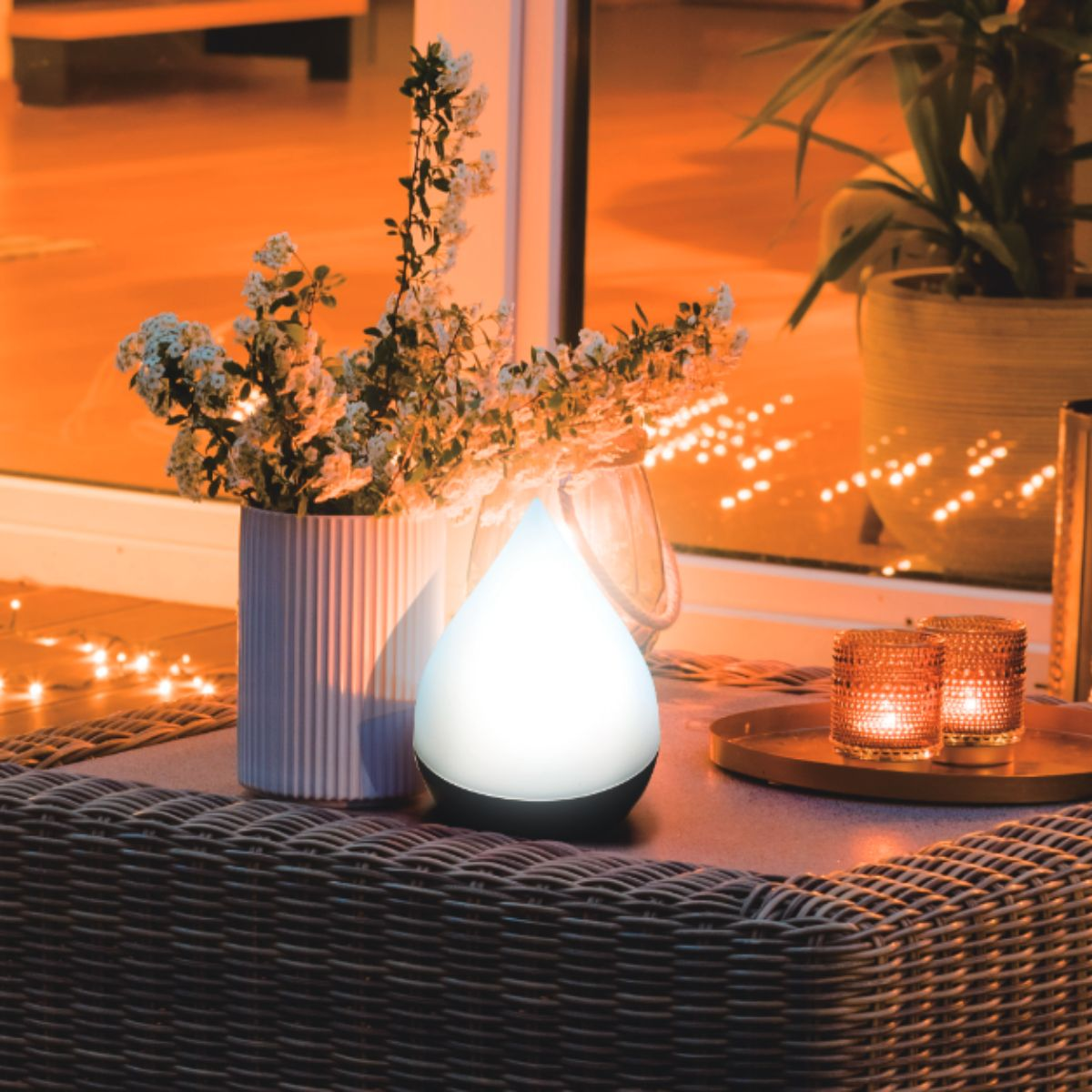 A tear-shaped lamp lights up a table with flowers and candles, with a window reflection.