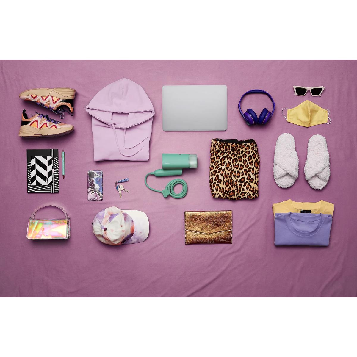 Eine lilafarbene Decke mit Kleidung und Accessoires: Schuhe, Hoodie, Laptop, Kopfhörer, Maske, Haartrockner, Rock, Hausschuhe, Tasche, Kappe, Clutch, T-Shirts.