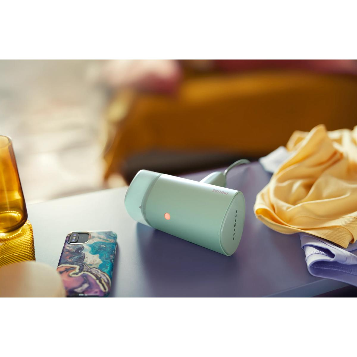 Ein hellgrüner Handdampfer liegt auf einem lila Tisch neben einem Handy und Kleidung.
