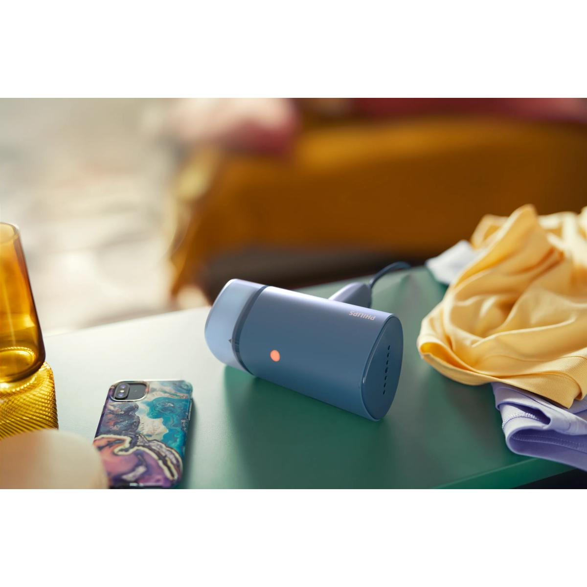 Een blauwe handstomer met oranje lampje staat op een groene tafel naast een telefoon en kleding.
