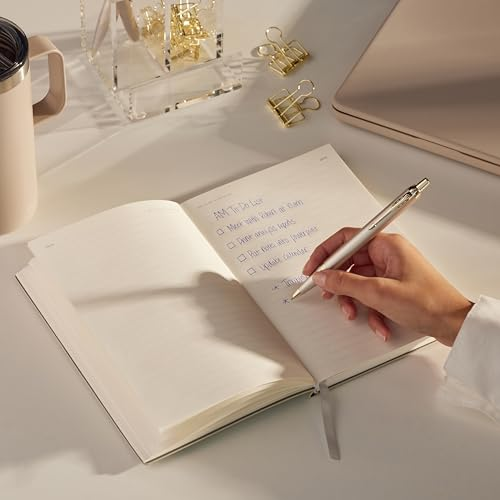 Eine Hand schreibt mit einem silbernen Stift in ein Notizbuch. Goldene Büroklammern und eine Tasse sind in der Nähe.