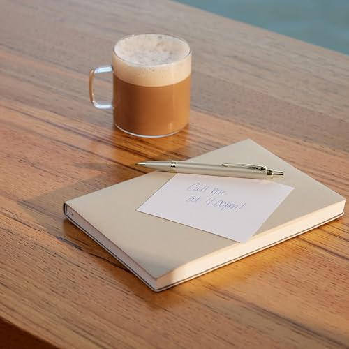 Eine Kaffeetasse aus Glas steht auf einem Holztisch neben einem Notizbuch und Stift.