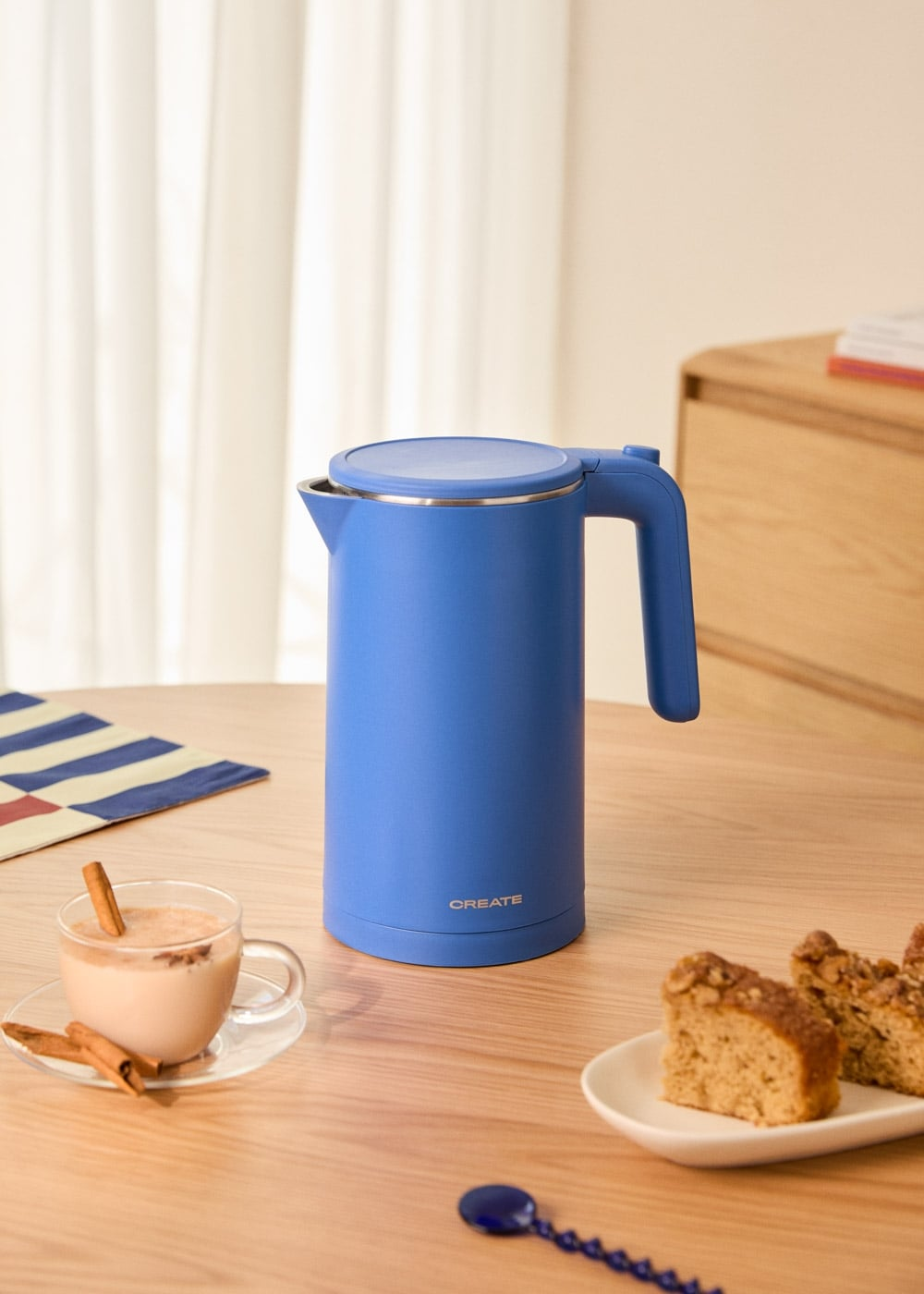 Un hervidor azul está sobre una mesa de madera junto a una taza de café y pastel.