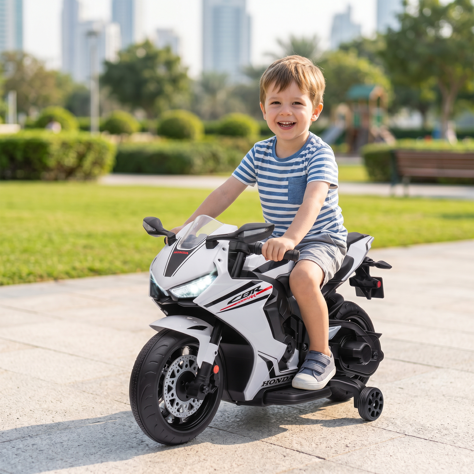 Un niño sonriente monta una motocicleta de juguete blanca en un parque con edificios detrás.