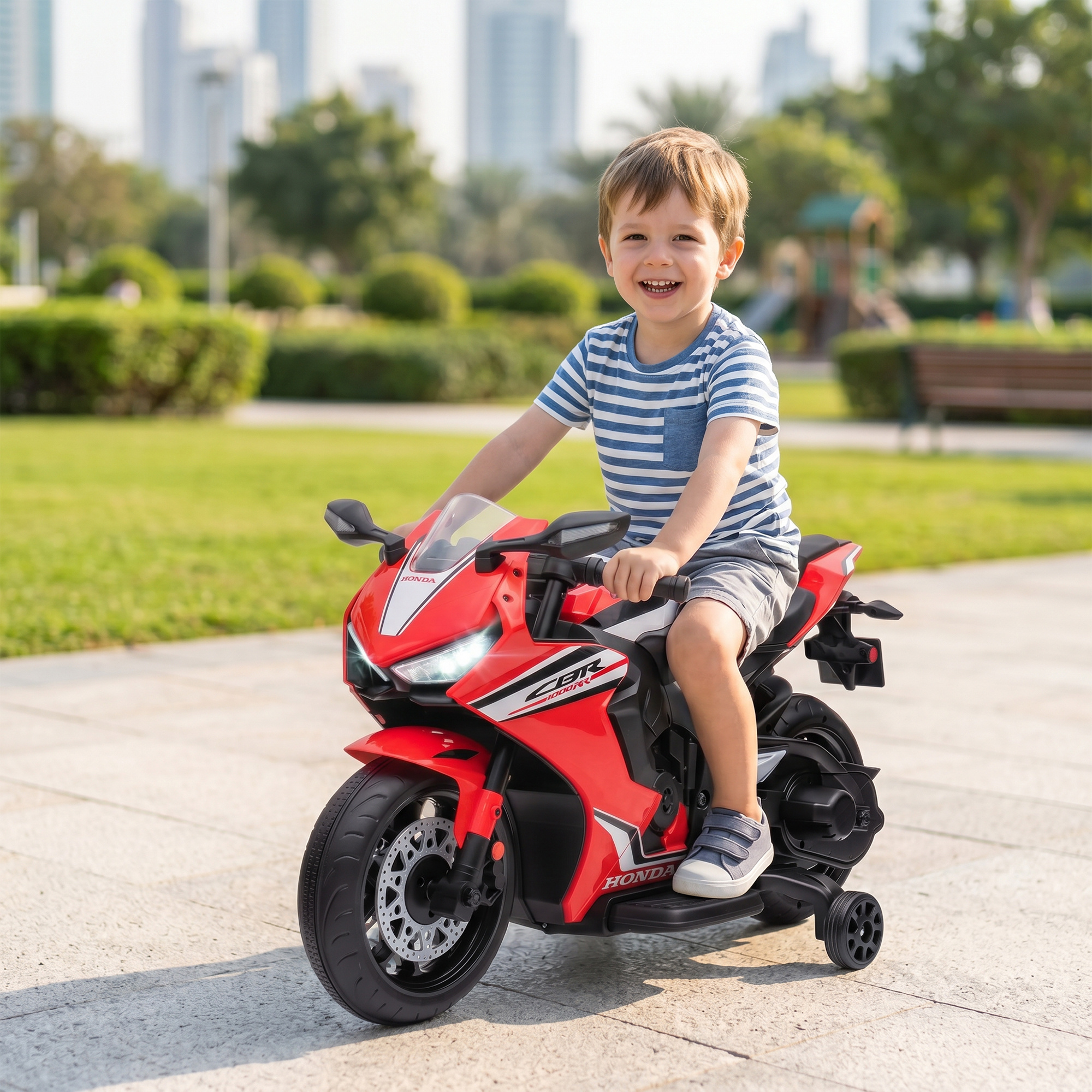 Un niño sonriente monta una motocicleta de juguete roja en un parque con edificios al fondo.