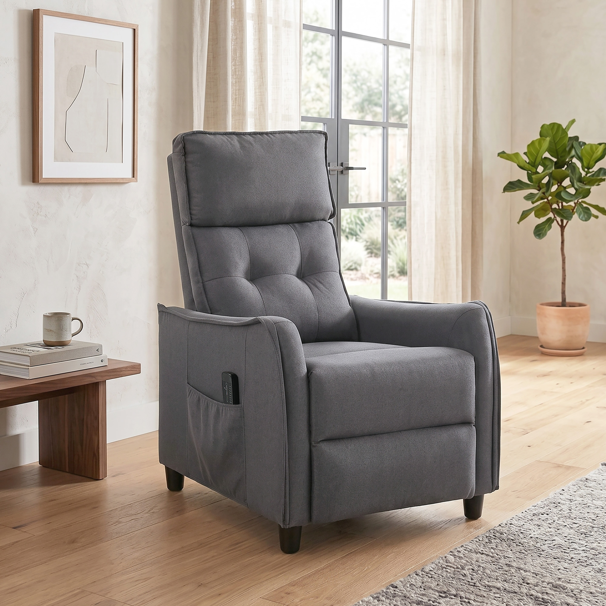 Un sillón reclinable gris está sobre un suelo de madera junto a una mesa con libros y una taza.