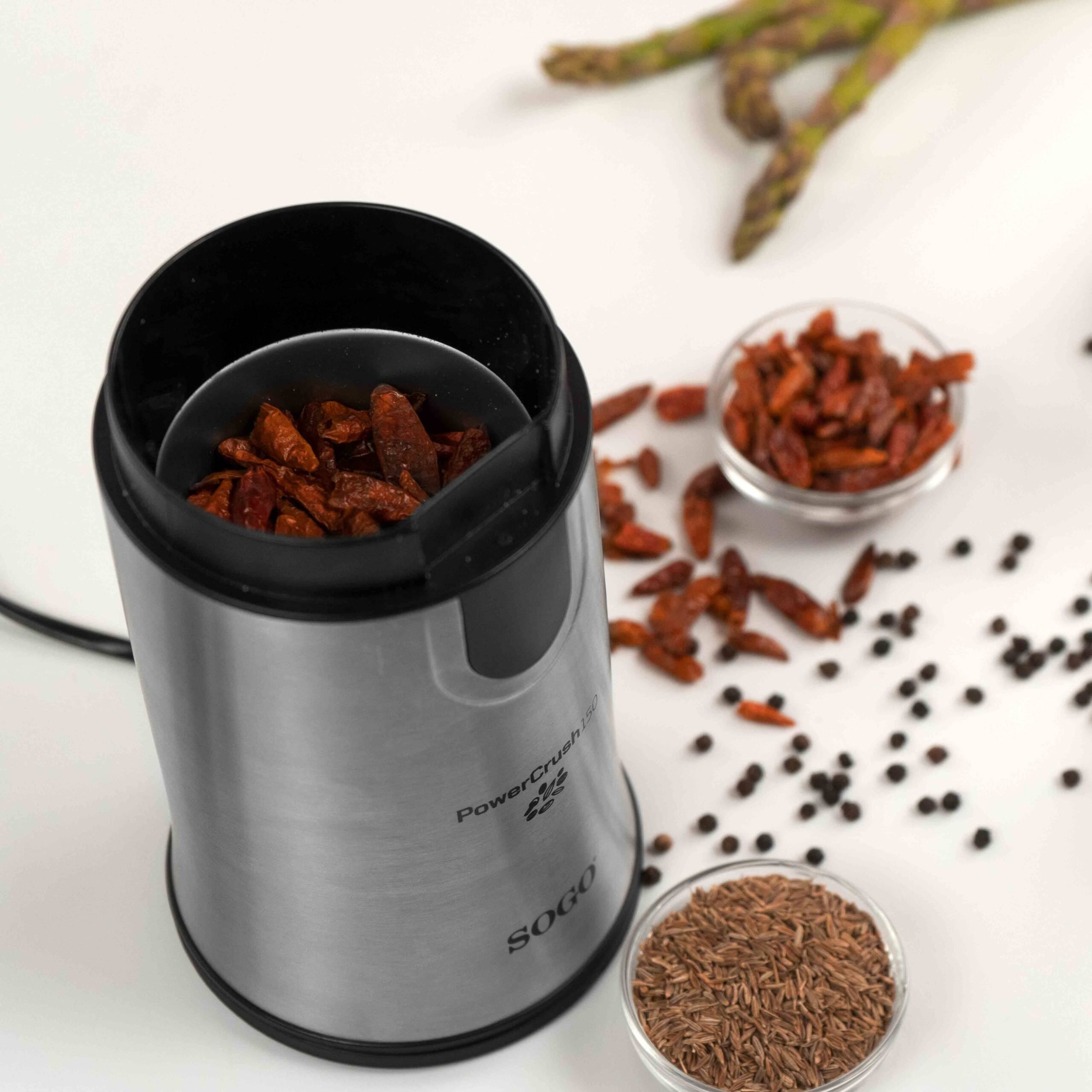 Un molinillo eléctrico plateado con chiles secos dentro, junto a cuencos de chiles, granos de pimienta y comino.