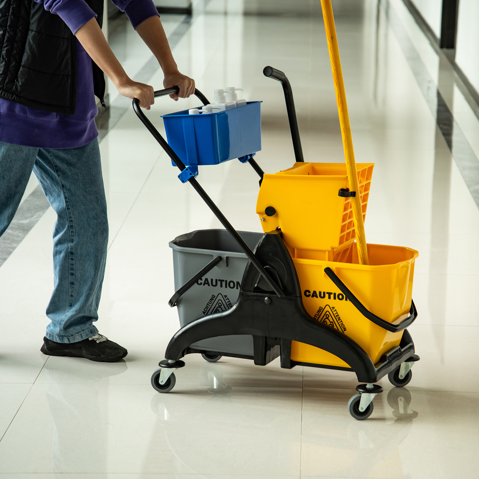 Una persona empuja un carrito de limpieza con cubos amarillos y grises en un pasillo.