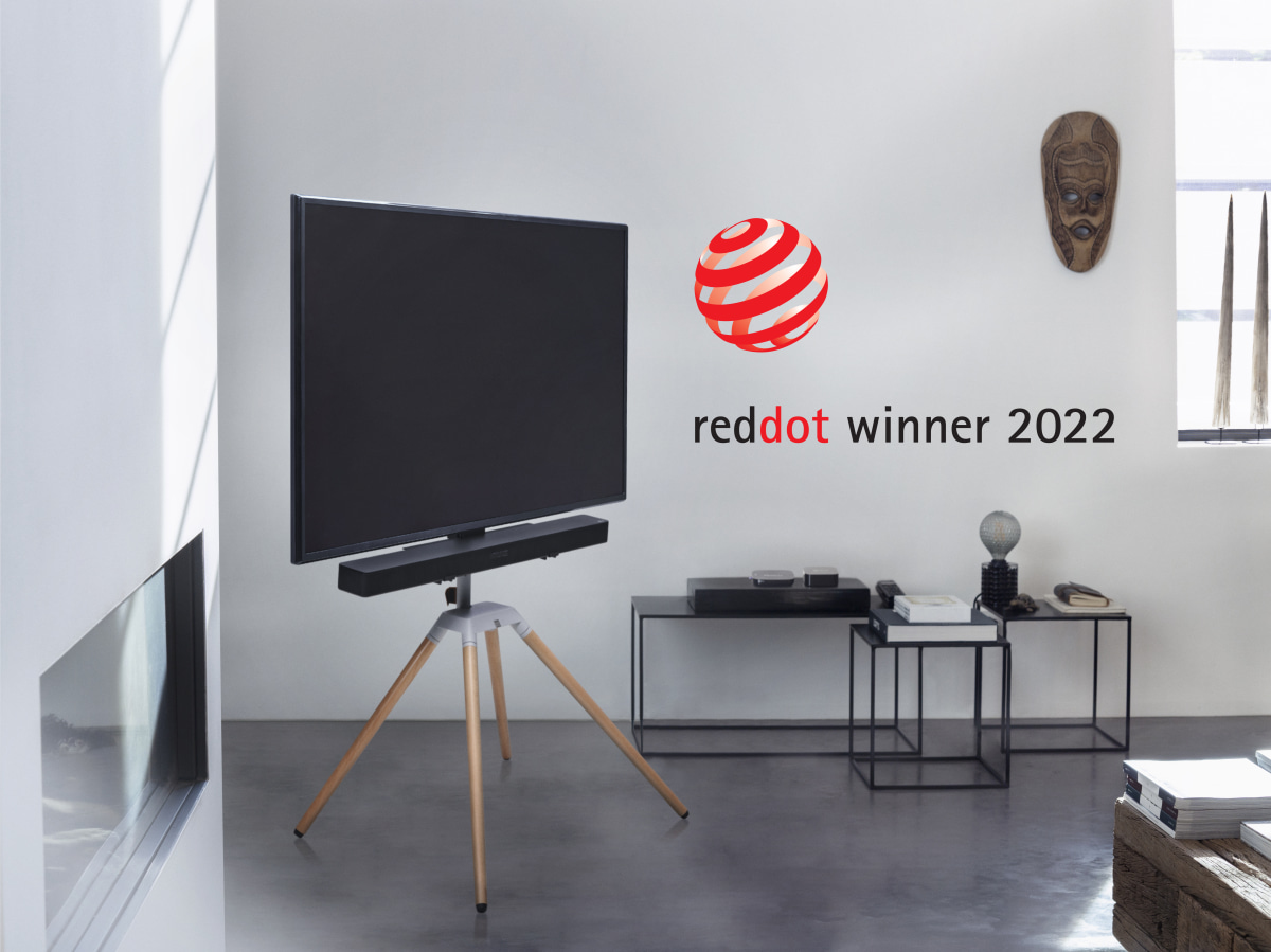 Ein Fernseher auf einem Dreibeinstativ mit Soundbar, Red Dot-Logo und Wanddekoration.