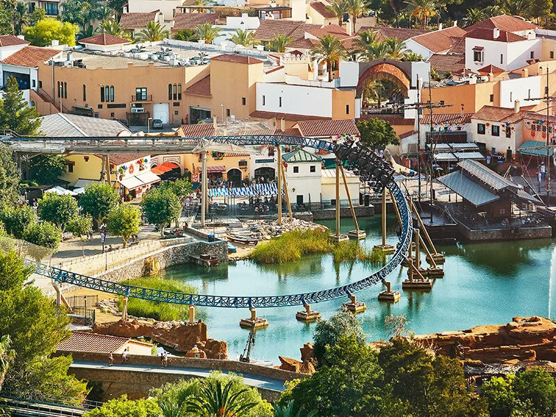 Una montaña rusa cruza un lago con edificios de estilo mediterráneo y barcos.