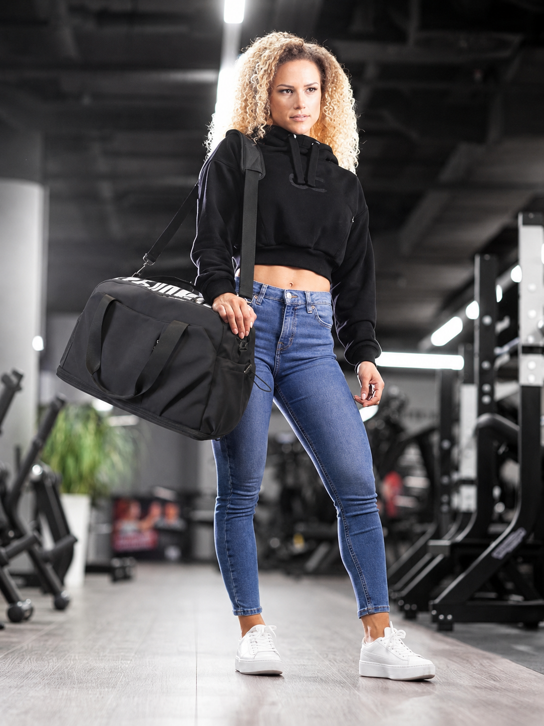 Frau mit lockigem Haar trägt Crop-Hoodie, Jeans und Sporttasche im Fitnessstudio.