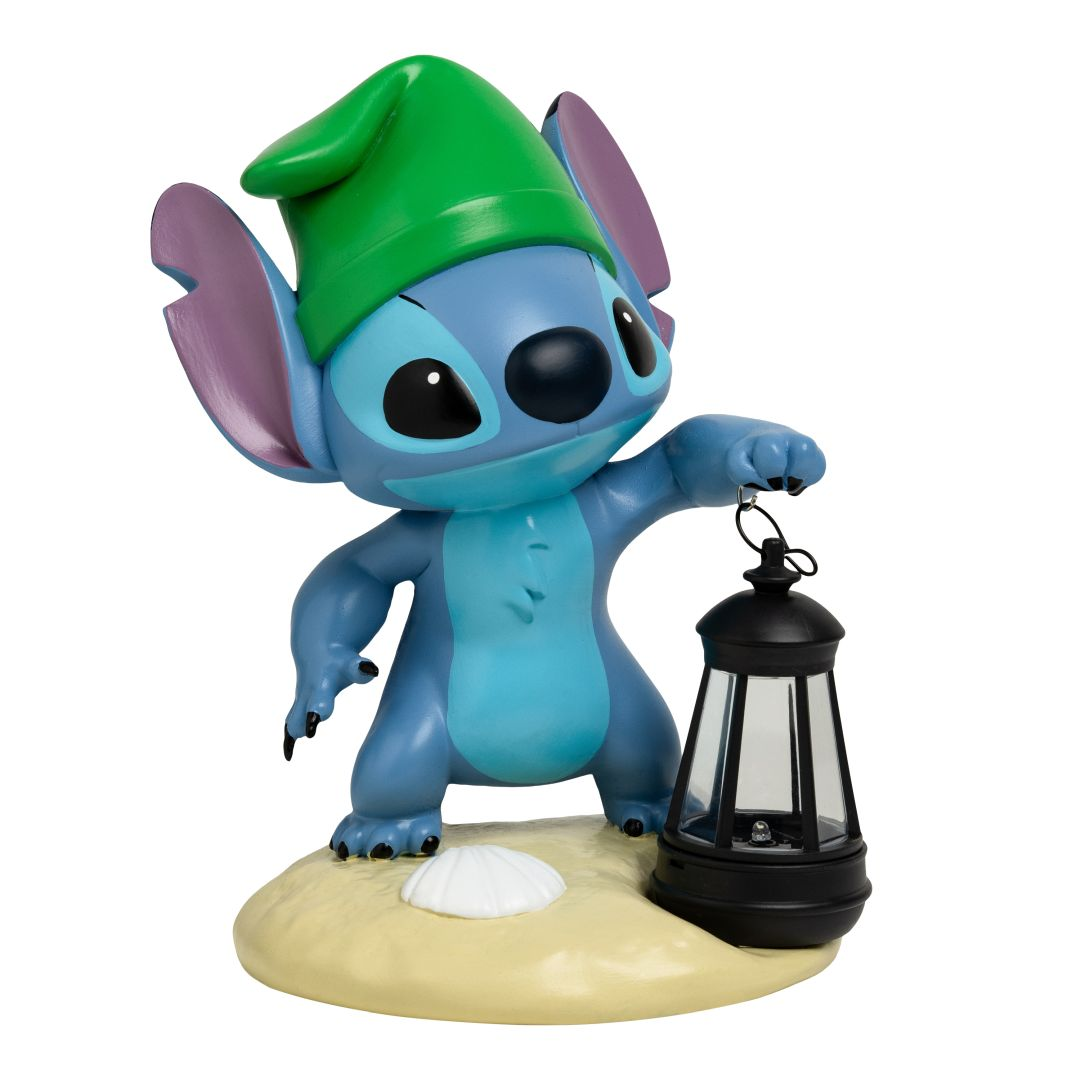 Stitch w zielonej czapce trzyma latarnię na piaszczystym podłożu z muszlą.