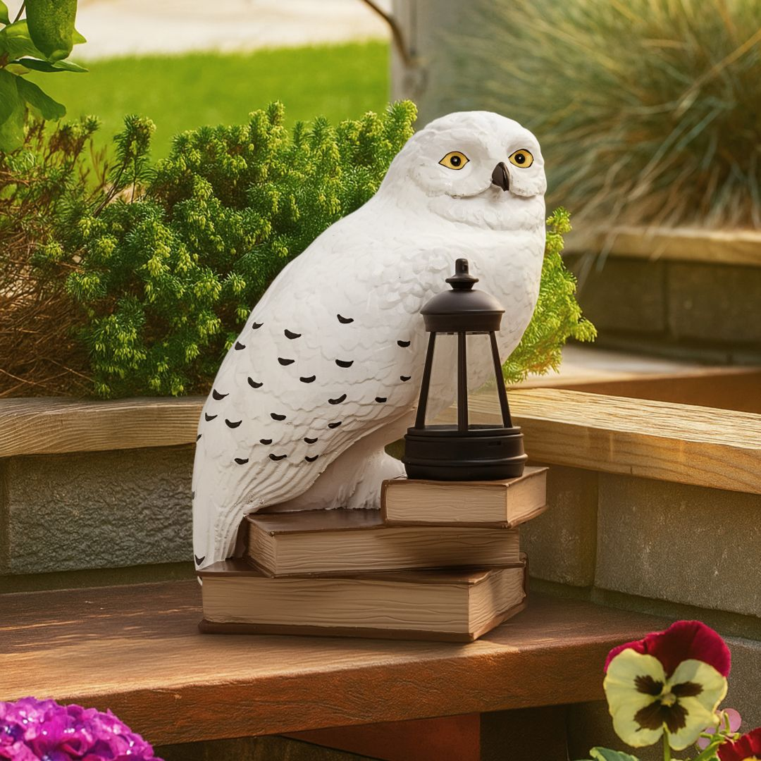 Una estatua de búho blanco se sienta sobre libros apilados junto a un farol, con plantas y flores.
