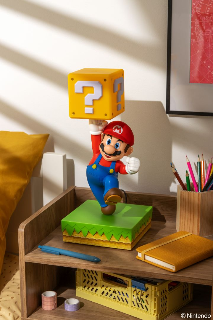 Figurka Mario trzyma żółty blok z pytajnikiem nad głową, na zielonym postumencie.