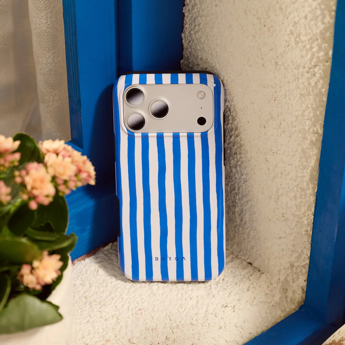 A blue and white striped iPhone case rests on a textured surface beside pink flowers.