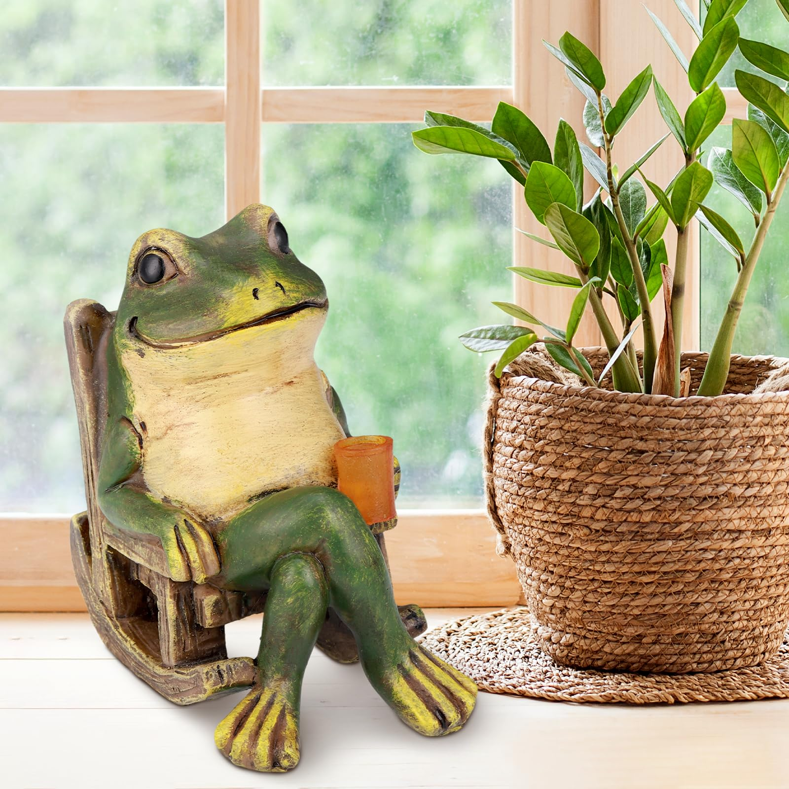 Grüne Froschfigur sitzt in Schaukelstuhl mit Getränk neben Pflanze.