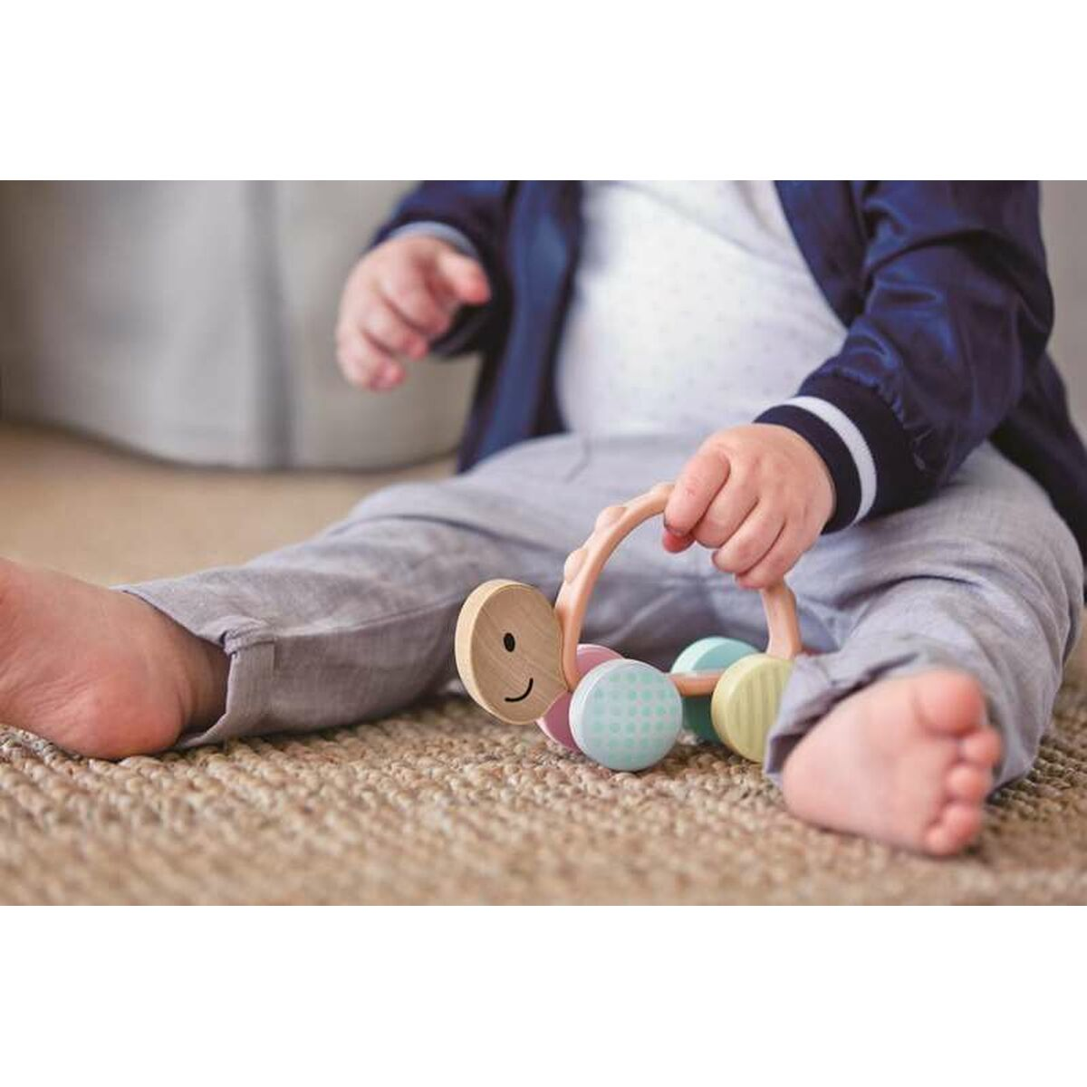 Ein Baby sitzt auf einem Teppich und hält ein Holzrasselspielzeug in Form einer Raupe mit bunten Rädern.