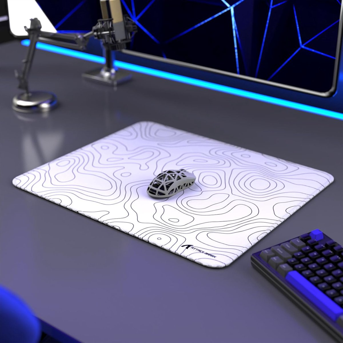Gray mouse on a white topographic pattern mousepad. Keyboard and monitor in the background.