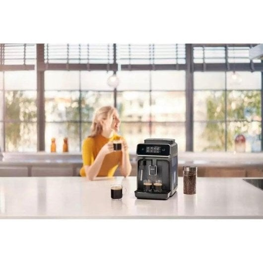 Een vrouw in een geel shirt houdt een kop koffie vast, met een koffiezetapparaat en bonen.