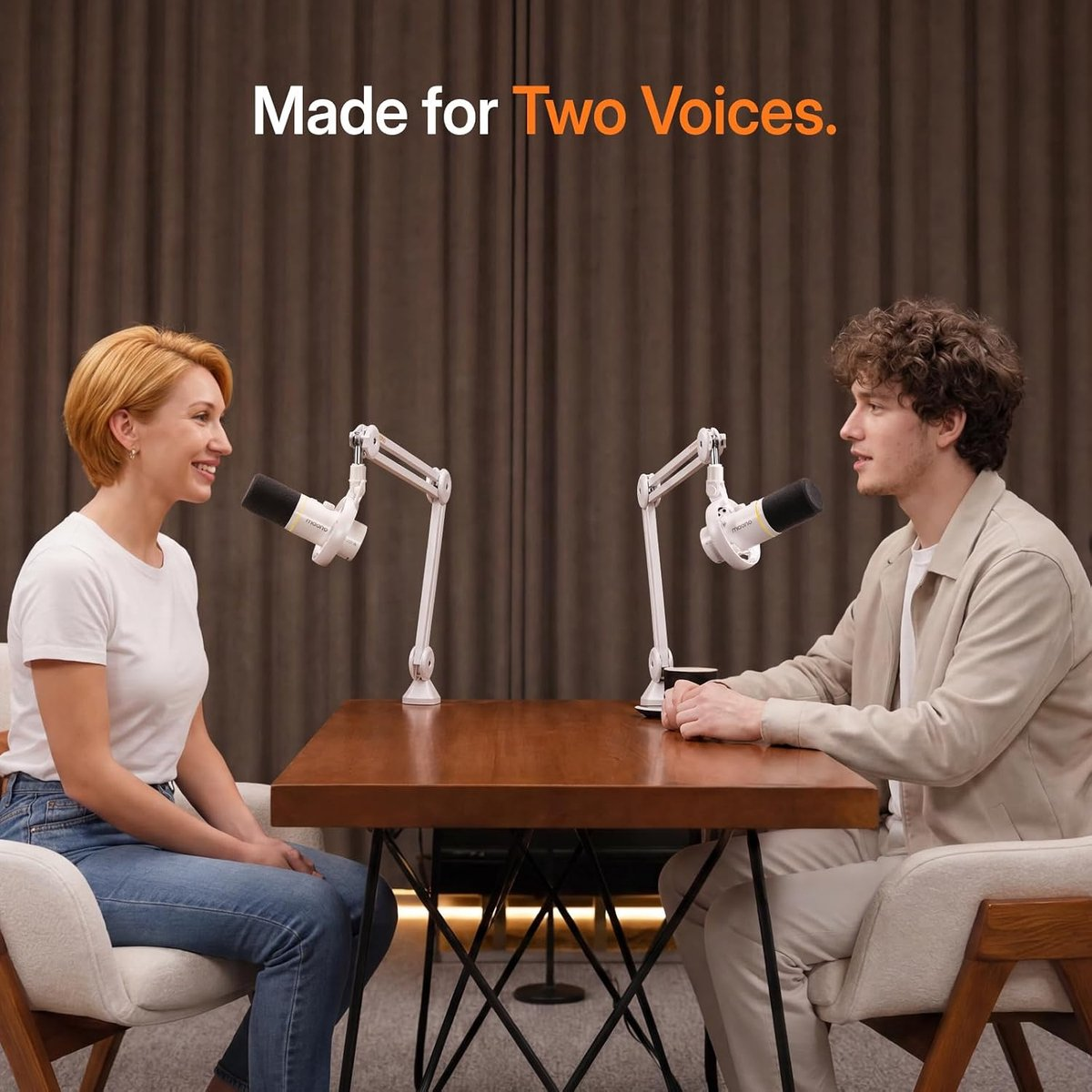 Two people sit at a wooden table with microphones in front of them.