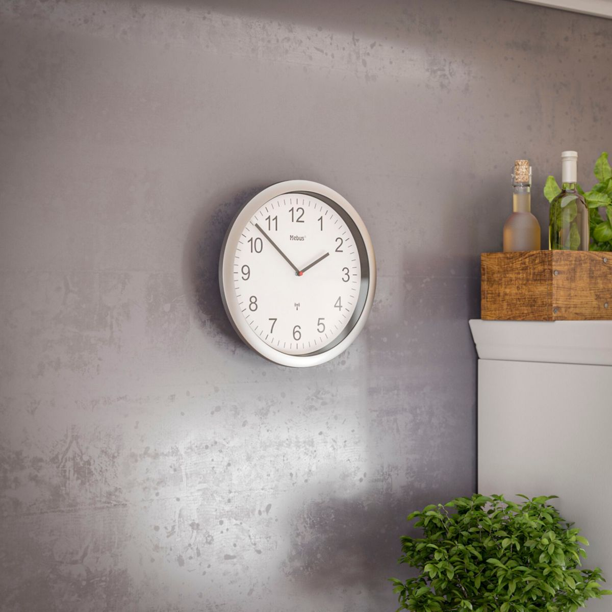 Un orologio Mebus bianco con numeri neri è appeso a un muro grigio. Sotto c'è una pianta.
