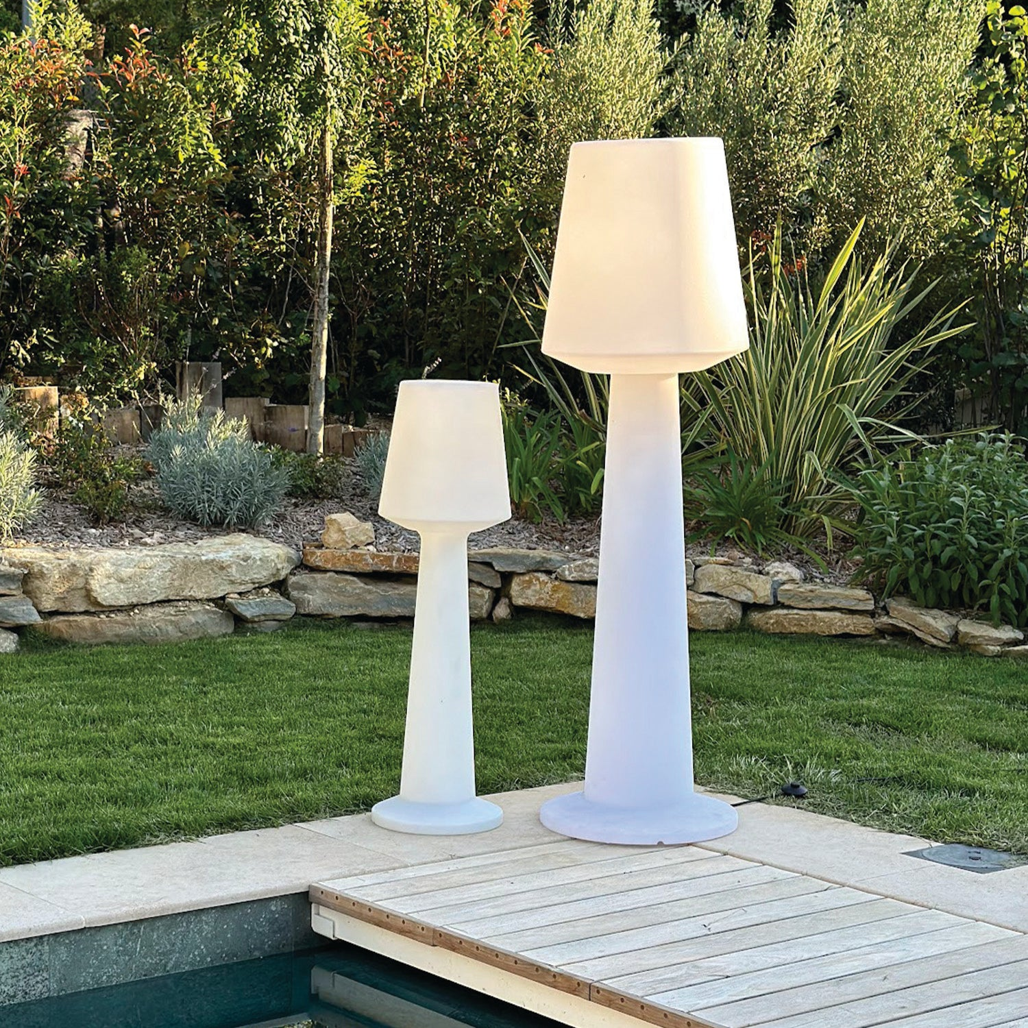 Zwei weiße Stehlampen stehen auf einer Holzterrasse neben einem Pool, im Hintergrund grünes Gras.